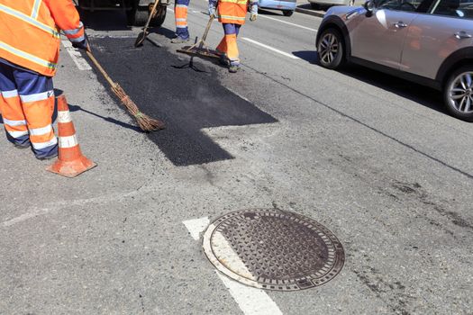 The working group of road workers near the manhole repairs part of the road with fresh hot asphalt and smoothes it with various hand tools - a shovel, level, broom.