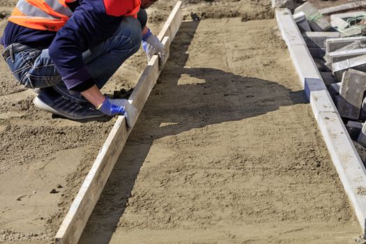 On the sidewalk, a worker cleans and levels the sandy platform with a wooden board, preparing the foundation for laying the paving slabs.