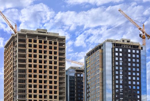 Fragment of the facade of a modern building under construction from glass and concrete, reflection of the blue cloudy sky in the glass walls of buildings, tower construction cranes are directed upwards.