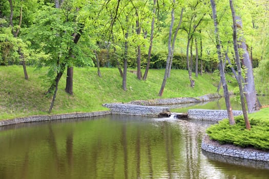 A forest lake with a cascade overflow of water in a beautiful park is located among various deciduous trees, framed on the banks of connected cobblestones, green grass and clipped shrubs under soft sunlight.