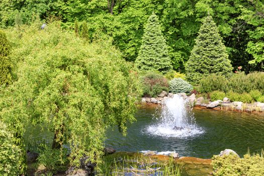 A small fountain in a decorative pond, surrounded by trees and bushes in a beautiful park, refreshes the air in hot summer in the rays of soft sunlight.