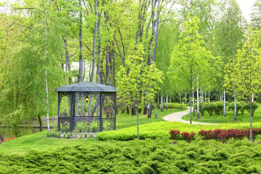 An openwork metal gazebo with a soft lamp in the center stands on a green lawn with decorative flowers, near a paved path passing by, framed on all sides by bushes and lanterns under soft sunlight.