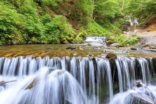 Daytime soft light in the forest illuminates the clear water of a mountain river in a cascade waterfall in the Carpathians against the background of bright green foliage.