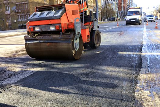 A heavy vibratory roller compacts hot asphalt on a repaired asphalt surface in the middle of the roadway on a clear sunny day.