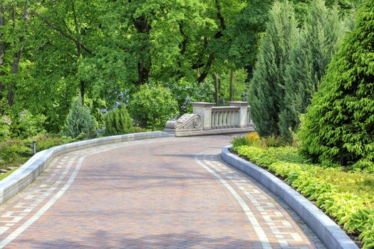 A laid flat path in a beautiful park passes through a green lawn with decorative flowers, framed on both sides with various bushes, well-kept trees and lanterns under soft sunlight.
