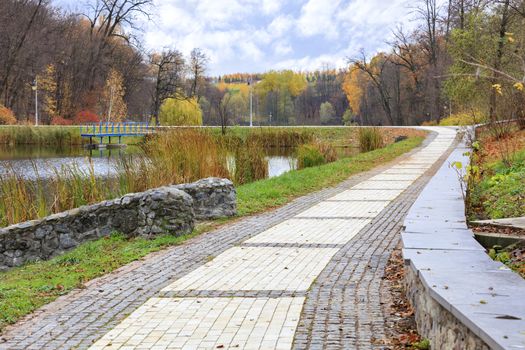 The road of paving slabs framed in cobblestone rounds the city pond in the autumn park.