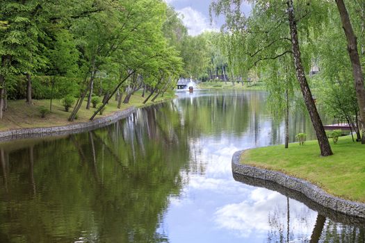A forest pond with fountain in a beautiful picturesque summer park is located among various deciduous trees, framed on the banks of connected cobblestones and green grass.