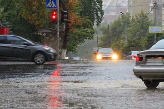 Torrential rain waters passing cars at the crossroads of the road and the red light of the traffic light is reflected in the flow of water on a city street.