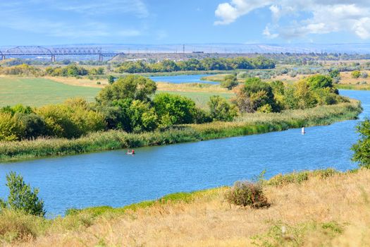 The wide bend of the Southern Bug River is immersed in the greenery of the garden and dried grass on the slope in the foreground, the river reaches the railway bridge on the horizon.