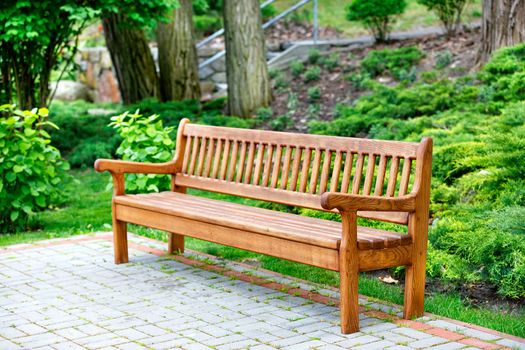 A beautiful and elegant handmade oak bench stands in a park in a recreation area on paving slabs with green bushes and large trees to the rear.