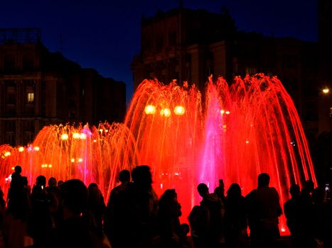 Dark silhouettes of people around the fountainin the late evening, red neon lights illuminate the jets of a powerful urban singing fountain.