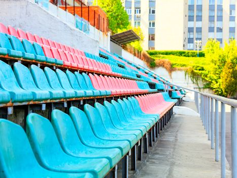 The rhythmic rows of empty turquoise and red plastic chairs of an old sports stadium against the background of sunlight in the background.