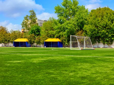 Green lawn of a football field with gates, netting and tents for team players on a summer day against a background of green foliage and a slightly cloudy blue sky.