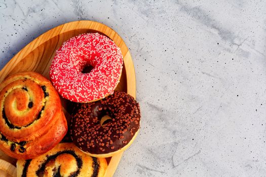 Delicious donuts with chocolate and strawberry icing and buns with poppy seeds and freshly baked raisins, close-up on an oval wooden tray and a gray concrete surface. Pastry sweets for breakfast, image with copy space.