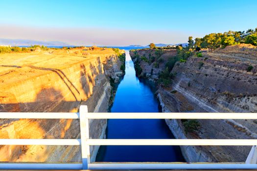 The Corinth Canal on a morning summer day illuminates the bright rising sun of Greece, a view of the Gulf of Corinth from the height of a pedestrian bridge.