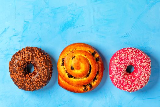Delicious donuts with chocolate and strawberry icing and buns with and freshly baked raisins, sprinkled with coconut and fruit chips on top, close-up on a light turquoise surface. Pastry sweets for breakfast, image with copy space.