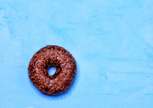 A delicious donut with chocolate icing sprinkled with fruit chips on top, close-up on a blue concrete surface with cold light. Pastry sweets for breakfast, image with copy space.