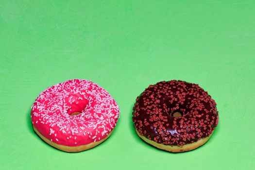 Two delicious donuts with chocolate and strawberry icing sprinkled with coconut and fruit chips on top, close-up on a green rough surface. Pastry sweets for breakfast, image with copy space.