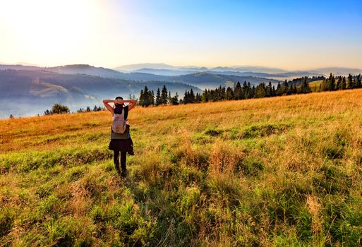 A young woman breathes the aroma of wild herbs and flowers early in the morning in the rays of the rising sun, meets dawn on a hill meadow and looks at the foggy Carpathian mountains.