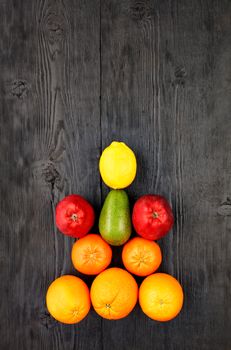 Ripe oranges, tangerines, apples, avocados and lemon lie symmetrically on an old black wooden surface, flat lay, image with copy space, in vertical image.