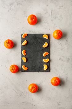 Pieces of tangerine, bright and juicy, lie on a gray slate surrounded by whole orange tangerines located on a gray concrete background, copy space, vertical image.