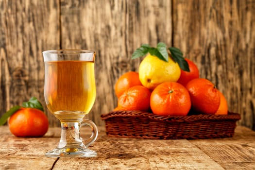 Citrus tea in a glass cup, warm healthy drink in the cool seasonon, background of mandarin and lemon fruits piled in a wicker basket on an old wooden table in blur, copy space.