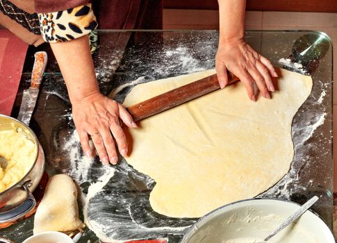 Housewife hands rolls the dough with a wooden rolling pin and prepares the ingredients for modeling dumplings in Ukrainian national cuisine.