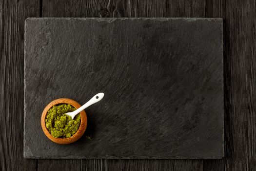 Matcha green tea powder in oak cup with white ceramic spoon with kitchen cutting board made of black slate, on wooden dark background, flat lay, copy space.