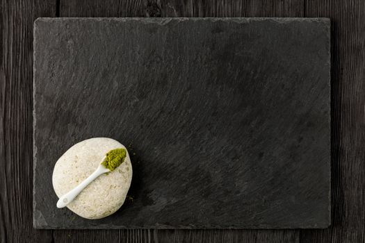 Matcha green tea powder in white ceramic spoon on white stone with kitchen cutting board made of black slate on wooden dark background, flat lay, copy space.