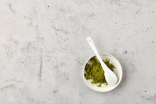 Matcha green tea powder in white ceramic cup with ceramic spoon on gray concrete background, flat lay, copy space.