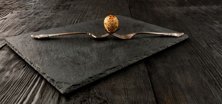 Quail egg on two old crossed forks on a kitchen cutting board in black of slate and a dark wooden background. Low key. Angled view.