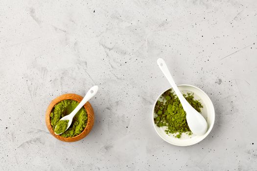 Matcha green tea powder in oak cup with white ceramic spoon and in ceramic cup with white ceramic spoon on gray concrete background, flat lay, copy space.