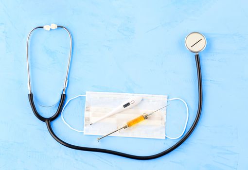 Stethoscope, medical mask, electronic thermometer and syringe with a yellow vaccine on a blue concrete background. The concept of protection against viruses, influenza and high body temperature. Copy space flat lay.