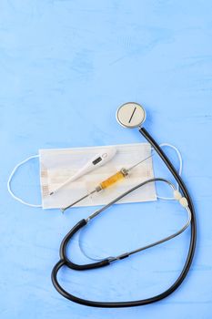 Stethoscope, medical mask, electronic thermometer and syringe with a yellow vaccine on a blue concrete background. The concept of protection against viruses, influenza and high body temperature. Copy space, vertical image.