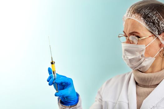 A doctor in a latex medical glove and a mask on his face checks the vaccine from a small syringe for injection. Virus protection concept. Medical theme on a light turquoise background.