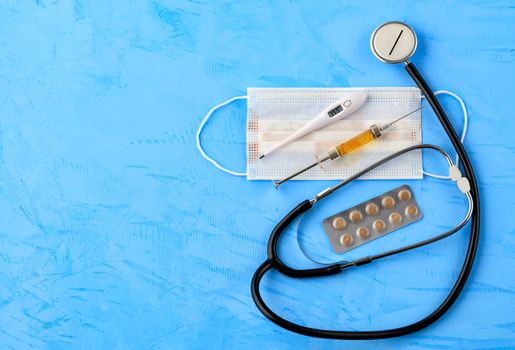 Stethoscope, medical mask, electronic thermometer and syringe with a yellow vaccine on a blue concrete background. The concept of protection against viruses, influenza and high body temperature. Copy space, top view.