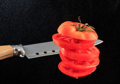 Flying red sliced tomato with a knife blade on a background of water dust in the backlight of a black background. Copy space, horizontal image.
