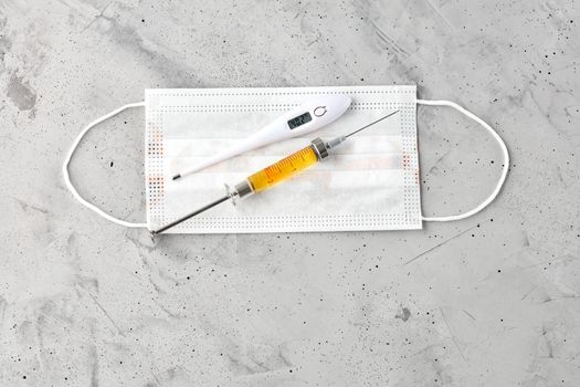 Medical mask, electronic thermometer and syringe with a yellow vaccine on a gray concrete background. The concept of protection against viruses, influenza and colds. Copy space, top view.
