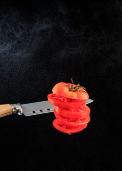 Flying red sliced tomato with a knife blade on a background of water dust in the backlight of a black background. Copy space, vertical image.