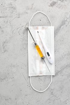 Medical mask, electronic thermometer and syringe with a yellow vaccine on a gray concrete background. The concept of protection against viruses, influenza and colds. Copy space, vertical image.