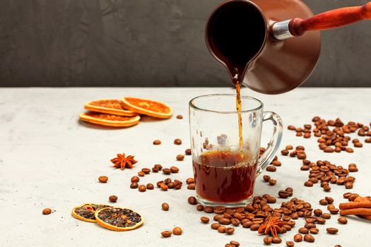 Freshly brewed espresso coffee is poured from a cezve into a transparent glass cup, surrounded by coffee beans, dried orange and lemon slices, anise stars and cinnamon sticks on a gray concrete background.