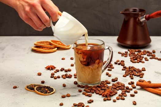 In a freshly made espresso in a transparent glass cup, a man's hand adds white cream, surrounded by coffee beans, dried slices of orange and lemon, anise stars and cinnamon sticks on a gray concrete background.