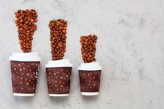 Coffee beans and three disposable cardboard mugs of different capacities for hot drinks, with a lid on a gray concrete background.