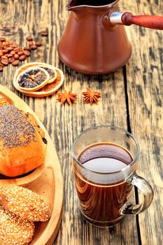 Freshly made espresso coffee from Turks is poured into a transparent glass cup against an old wooden table with coffee beans, dried orange and lemon, anise, cinnamon and rolls.