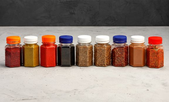 Jars glass transparent with various asian spices ground paprika, corinade, hops, zira, sumac, caraway seeds, cinnamon, saffron, allspice on a gray concrete background.