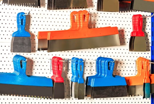 Steel spatulas and scrapers of various shapes and purposes, serrated and polished, with plastic handles hang on a metal perforated white stand.