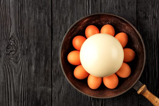 A large ostrich egg surrounded by chicken eggs in an old cast-iron pan that stands on an old black wooden surface, low key image, top view, copy space.