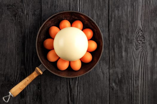 Ostrich egg surrounded by chicken eggs in an old cast-iron skillet, which is standing on an old black wooden surface, top view, copy space.