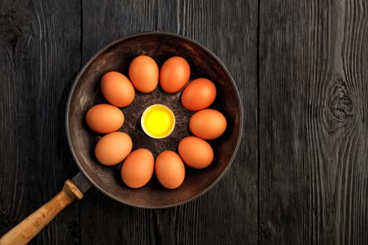 Bright chicken yolk of a broken egg surrounded by chicken eggs in an old cast-iron pan, which is standing on an old black wooden surface, top view, copy space.