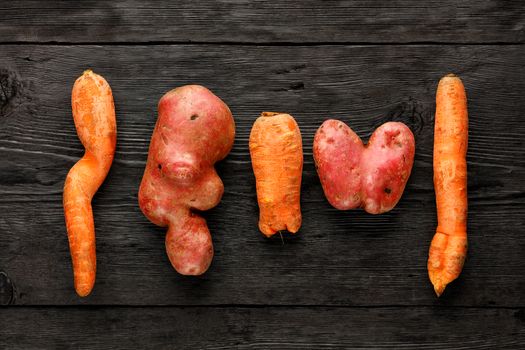 Ugly funny vegetables, heart-shaped potatoes and twisted carrot on a black wooden background. The concept of grungy vegetables or food waste. Top view, low key, copy space.